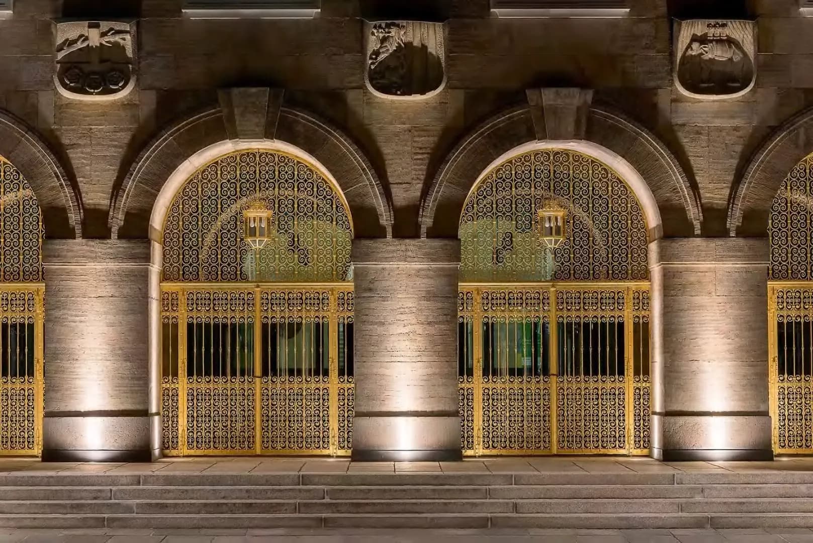 Stone facade and brass gates drawn out of the dark by ground-mounted uplights